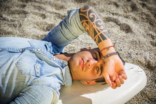 Stylish handsome young man laying on surfboard on beach in sunlight
