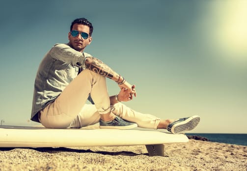 Stylish handsome young man in mirrored sunglasses sitting on surfboard on beach in sunlight