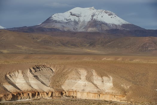Snow covered peak of the active Guallatiri volcano (6063 m) towering above the Altiplano and cliffs running along the valley of the River Lauca in Lauca National Park, northern Chile.