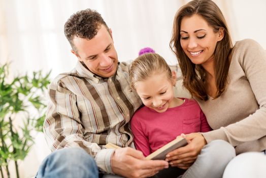 Beautiful young smiling family relaxing on sofa at living room while reading book.