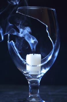 Extinguished candle with smoke inside broken wineglass on dark background