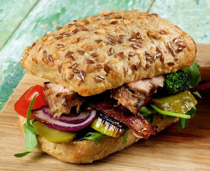 Delicious Homemade Burger with Roasted Pork, Marinated Cucumber, Stewed Vegetables, Greens and Red Onion on Brown Bread with Seeds closeup on Wooden background