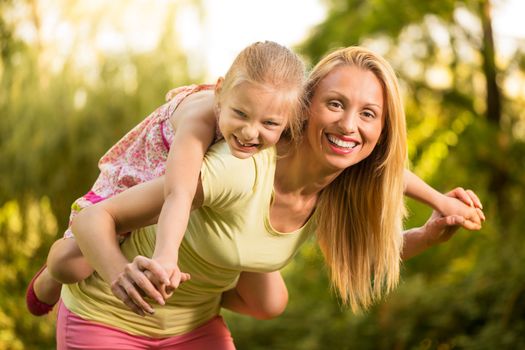 Beautiful mother giving her cute little girl piggyback and smiling in the park