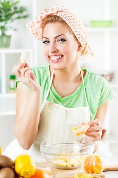 Beautiful young woman preparing orange for fruit salad in the kitchen. Looking at camera.