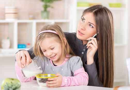 Overworked Business Woman and her little daughter in the morning. Overworked mother make phone calls before going to work. Daughter preparing cereal with milk for her daughter and spilling milk next to bowl.