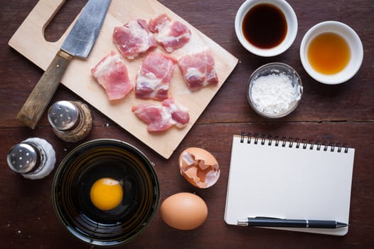 Preparation for cooking in home kitchen. Soft pork ribs on chopping board, notepad for check list, egg, and ingredients on wood table.
