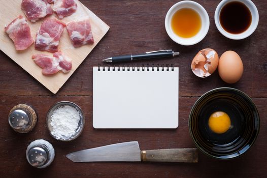 Preparation for cooking in home kitchen. Soft pork ribs on chopping board, notepad for check list, egg, and ingredients on wood table.
