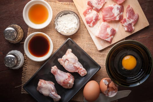 Preparation for cooking in home kitchen. Soft pork ribs on chopping board, chicken wings in ceramic dish, eggs, and ingredients on wood table.
