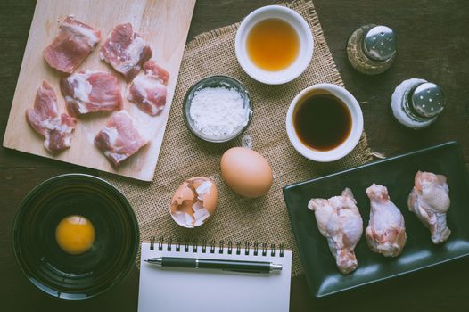 Preparation for cooking in home kitchen. Soft pork ribs on chopping board, chicken wings in ceramic dish, eggs, and ingredients on wood table with vintage filter effect.