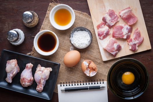 Preparation for cooking in home kitchen. Soft pork ribs on chopping board, chicken wings in ceramic dish, eggs, and ingredients on wood table.