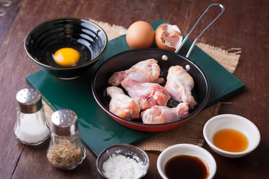 Preparation for cooking in home kitchen. chicken wings in small pan, eggs, and ingredients on wood table.