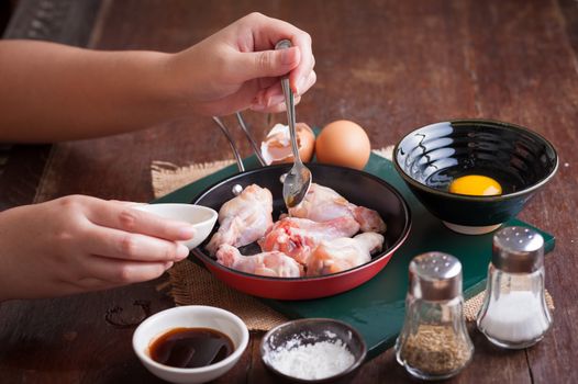 Preparation for cooking in home kitchen. Woman left hand pouring oyster sauce into fry pan to mix with chicken wings.