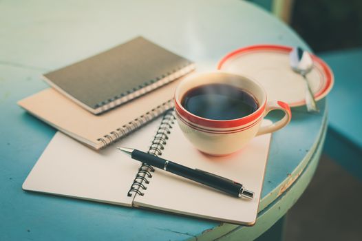 Coffee cup with notebooks and pen on blue round wood table in morning time with vintage filter effect