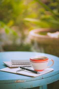 Coffee cup with notebooks and pen on blue round wood table in morning time with vintage filter effect