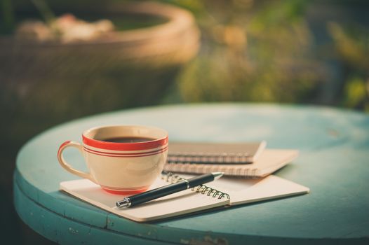Coffee cup with notebooks and pen on blue round wood table in morning time with film filter effect