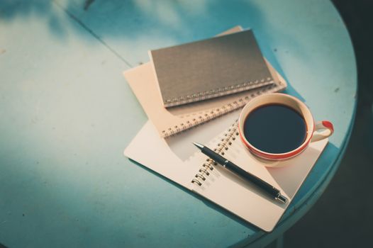 Coffee cup with notebooks and pen on blue round wood table in morning time with film filter effect