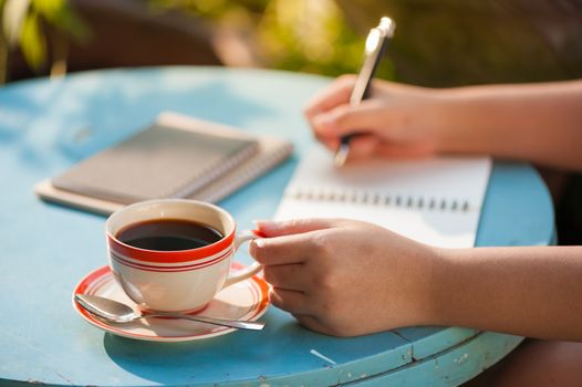 Woman left hand holding coffee cup while writing on notebook at outdoor area in cafe with morning scene
