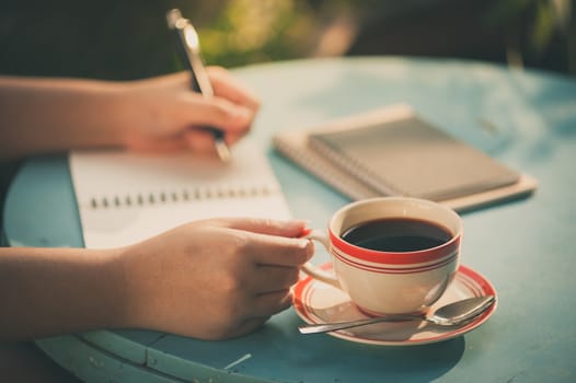 Woman right hand holding coffee cup while writing on notebook at outdoor area in cafe with vintage filter effect