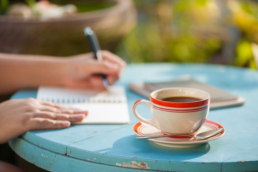 Coffee cup on wood table with woman hand writing on notebook in background at cafe with morning scene