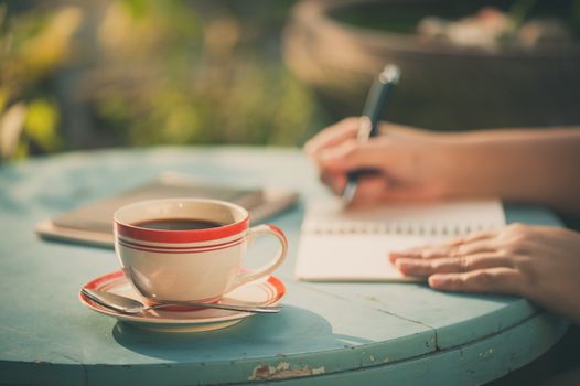 Morning scene of coffee cup on wood table with woman hand writing on notebook in background at cafe with vintage filter effect