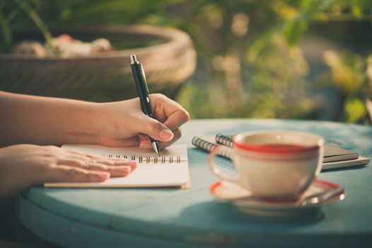 Woman left hand writing journal on small notebook in outdoor area at cafe with morning scene and vintage filter effect