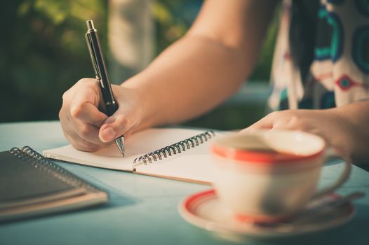 Woman hand writing journal on small notebook at outdoor area in cafe with morning scene and vintage filer effect