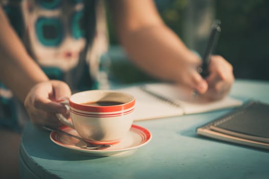 Morning scene of woman right hand holding coffee cup while writing on notebook in outdoor area at cafe with vintage filter effect