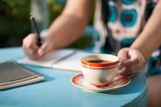 Woman left hand holding coffee cup while writing on notebook in outdoor area at cafe with morning scene