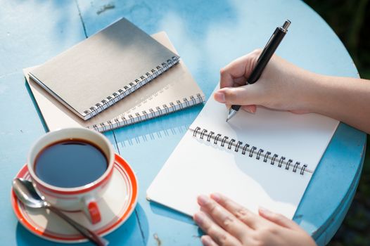 Woman right hand writing journal on small notebook in outdoor area at cafe with morning scene