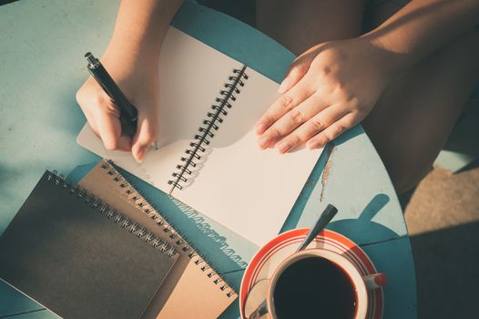 Woman right hand writing journal on small notebook in outdoor area at cafe with morning scene and vintage filter effect