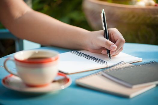 Woman left hand writing journal on small notebook in outdoor area at cafe with morning scene
