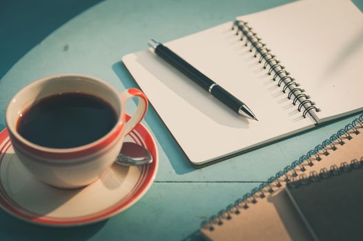 Small notebook and pen beside coffee cup on blue wood table at outdoor area in cafe with morning scene and vintage filter effect