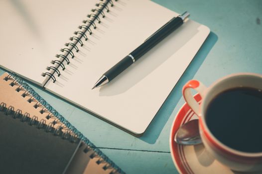 Small notebook and pen beside coffee cup on blue wood table at outdoor area in cafe with morning scene and vintage filter effect