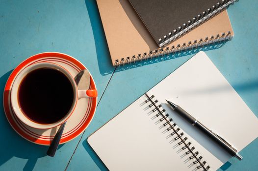 Small notebook and pen beside coffee cup on blue wood table at outdoor area in cafe with morning scene