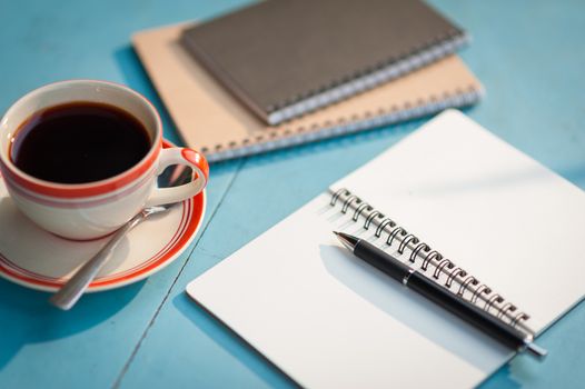 Small notebook and pen beside coffee cup on blue wood table at outdoor area in cafe with morning scene
