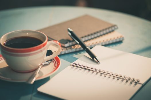 Morning scene of small notebook and pen beside coffee cup on blue wood table at outdoor area in cafe with vintage filter effect