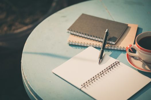 Small notebook and pen beside coffee cup on blue color round wood table at outdoor area in cafe with morning scene and vintage filter effect