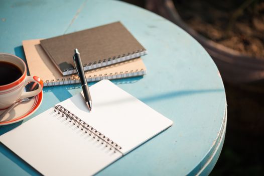 Small notebook and pen beside coffee cup on blue color round wood table at outdoor area in cafe with morning scene