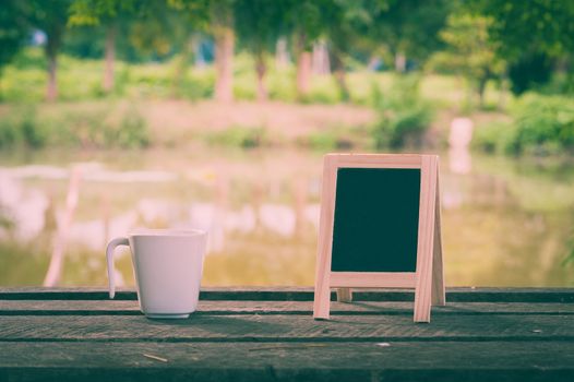 Small A-frame blackboard with blank area for text or message and coffee cup on rustic wood table in morning time with vintage filter effect