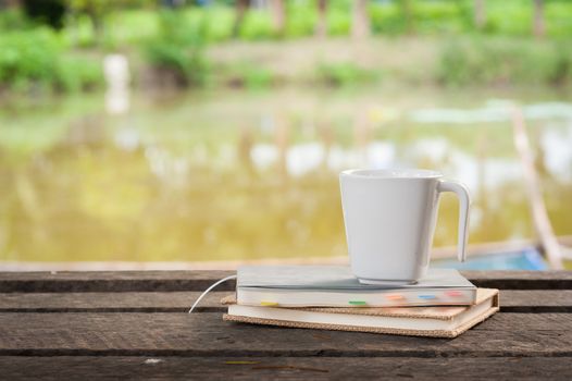 Coffee cup and notebooks on rustic wood table at outdoor area with rural lake view on blurry background in morning time