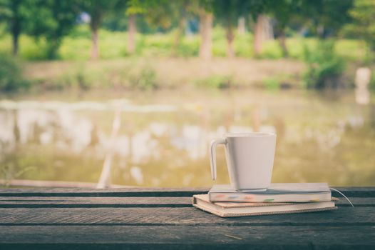Coffee cup and notebooks on rustic wood table at outdoor area with rural lake view on blurry background in morning time with vintage filter effect