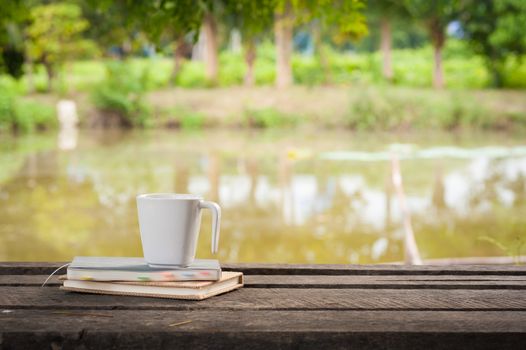 Coffee cup and notebooks on rustic wood table at outdoor area with rural lake view on blurry background in morning time