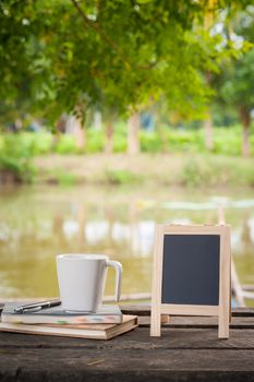 Small A-frame blackboard with blank area for text or message, notebooks and coffee cup on rustic wood table with rural lake view in blurry background at morning time