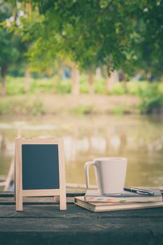 Small A-frame blackboard with blank area for text or message, notebooks and coffee cup on rustic wood table with rural lake view in blurry background at morning time with vintage filter effect