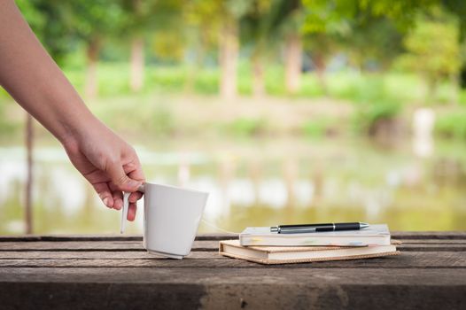 Woman left hand holding coffee cup beside notebooks on rustic wood bench with rural lake view in blurry background on weekend with morning scene