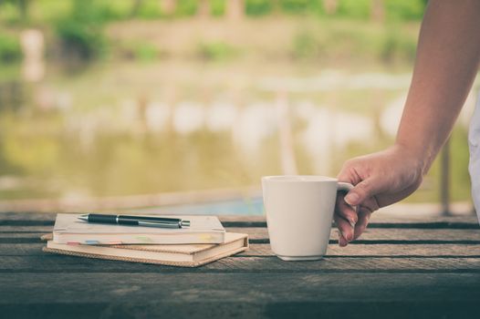 Woman hand holding coffee cup beside notebooks on rustic wood bench with rural lake view in blurry background on weekend with morning scene and vintage filter effect