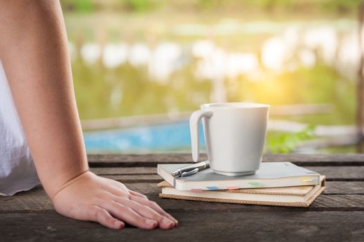 Woman hand put down beside coffee cup and notebooks on rustic wood bench with rural lake view in morning time on weekend