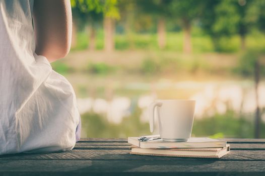Young woman sitting beside coffee cup and notebooks on rustic wood bench with rural lake view in morning time on weekend with vintage filter effect