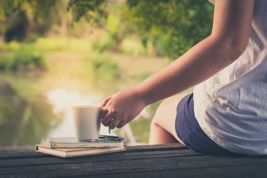 Woman left hand holding coffee cup beside notebooks on rustic wood bench with rural lake view in background in morning time on weekend with vintage filter effect