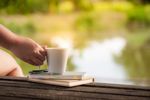 Woman right hand holding coffee cup beside notebooks on rustic wood bench in morning time on weekend